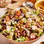 A large bowl of salad filled with mixed greens, cherry tomatoes, red onions, olives, chickpeas, croutons, and grated cheese. In the background, there's a small bowl of spices, a jar of peppers, and a wooden cutting board.