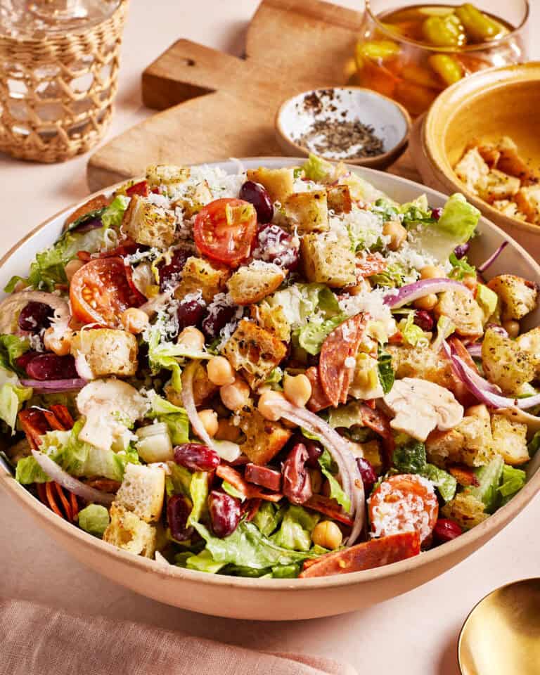 A large bowl of salad filled with mixed greens, cherry tomatoes, red onions, olives, chickpeas, croutons, and grated cheese. In the background, there's a small bowl of spices, a jar of peppers, and a wooden cutting board.