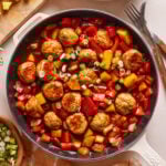 A skillet filled with sweet and sour meatballs, topped with chopped green onions. The dish also includes diced red bell peppers and pineapple chunks in a sauce. Plates, forks, and ingredients are around the skillet on a light-colored surface.