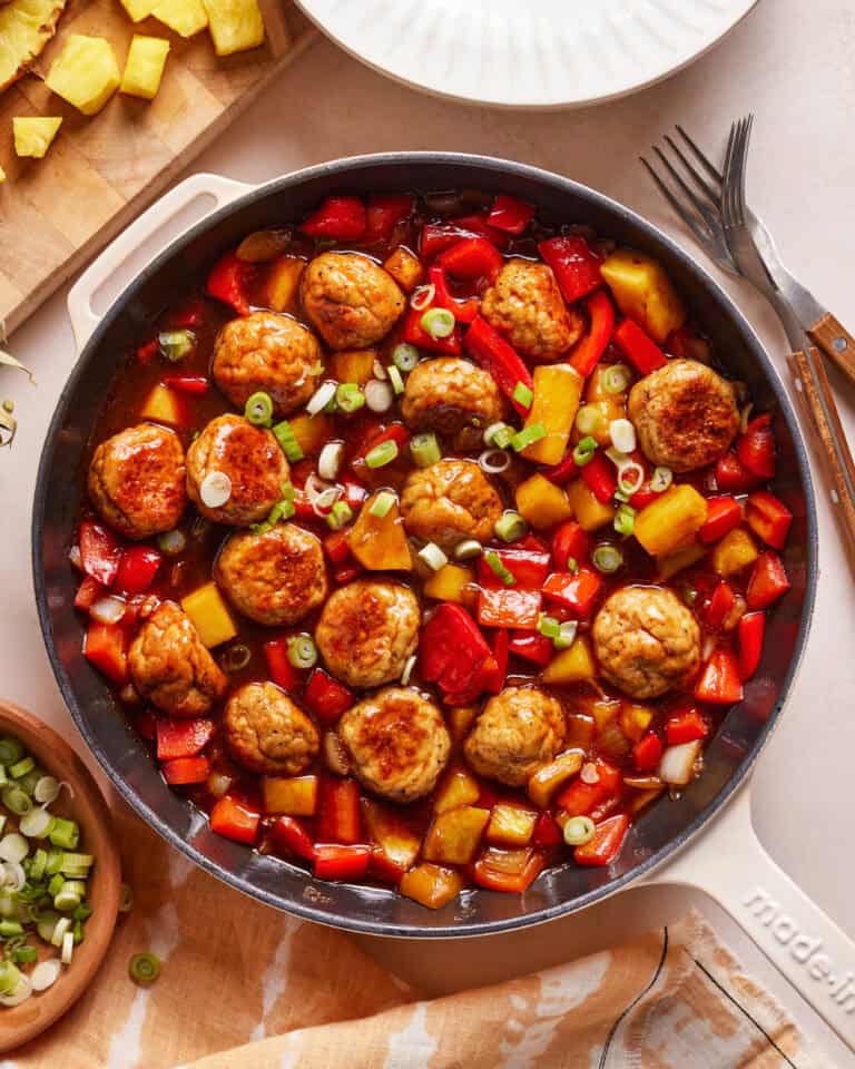 A skillet filled with sweet and sour meatballs, topped with chopped green onions. The dish also includes diced red bell peppers and pineapple chunks in a sauce. Plates, forks, and ingredients are around the skillet on a light-colored surface.