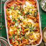 A baked pasta casserole in a white dish, topped with melted cheese, roasted zucchini slices, tomato sauce, and fresh basil, sits on a green tiled surface next to dishes, cutlery, and a bowl of shredded cheese.