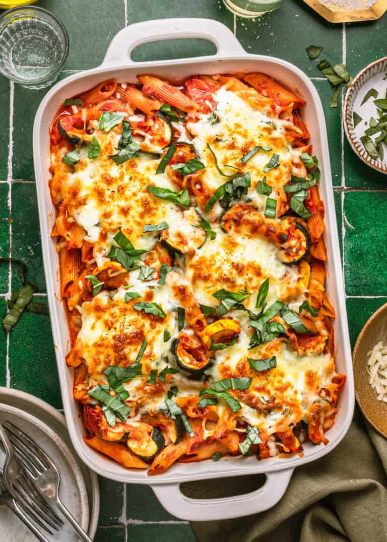 A baked pasta casserole in a white dish, topped with melted cheese, roasted zucchini slices, tomato sauce, and fresh basil, sits on a green tiled surface next to dishes, cutlery, and a bowl of shredded cheese.