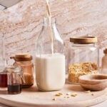Pouring plant-based milk into a glass bottle on a wooden surface. Surrounding jars contain sliced almonds, cinnamon, and vanilla extract. A small bowl filled with salt sits nearby, with a neutral-toned stone wall in the background.