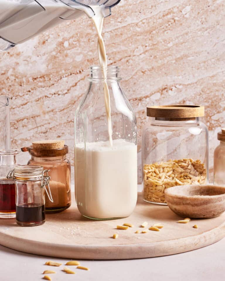 Pouring plant-based milk into a glass bottle on a wooden surface. Surrounding jars contain sliced almonds, cinnamon, and vanilla extract. A small bowl filled with salt sits nearby, with a neutral-toned stone wall in the background.
