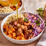 A bowl with quinoa, roasted sweet potatoes, spiced chicken, and coleslaw, with dressing being poured on top. Glasses of water and another bowl of food are in the background.