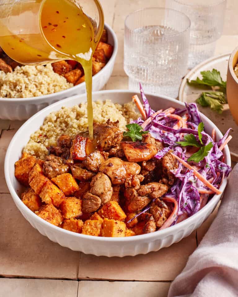 A bowl with quinoa, roasted sweet potatoes, spiced chicken, and coleslaw, with dressing being poured on top. Glasses of water and another bowl of food are in the background.