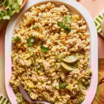 A platter of creamy pasta salad with fusilli, corn, and fresh cilantro. Lime wedges garnish the dish, and a serving spoon rests on the side. The table is set with green and white striped napkins.