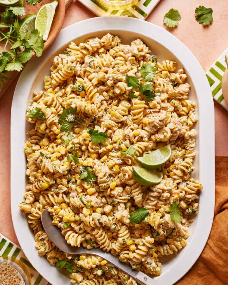 A platter of creamy pasta salad with fusilli, corn, and fresh cilantro. Lime wedges garnish the dish, and a serving spoon rests on the side. The table is set with green and white striped napkins.