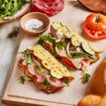 Two open-faced sandwiches with turkey, zucchini slices, fresh arugula, pickled onions, and tomato on flatbread, served on a wooden board with a small bowl of salt and pepper, arugula, and a glass of pickled onions nearby.
