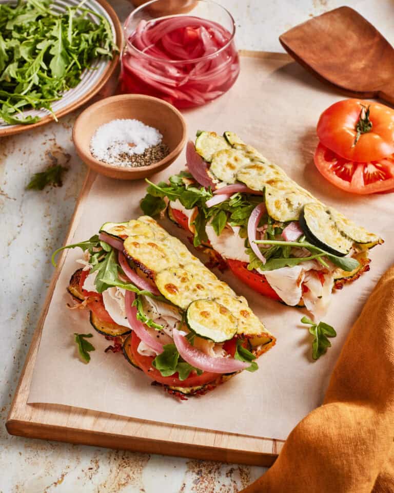 Two open-faced sandwiches with turkey, zucchini slices, fresh arugula, pickled onions, and tomato on flatbread, served on a wooden board with a small bowl of salt and pepper, arugula, and a glass of pickled onions nearby.