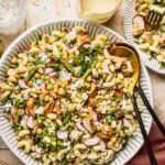 A large bowl of pasta salad with grilled chicken, radish slices, chickpeas, green peas, asparagus, herbs, and feta cheese. Two serving spoons rest in the bowl; a glass of dressing and a pink napkin are nearby.