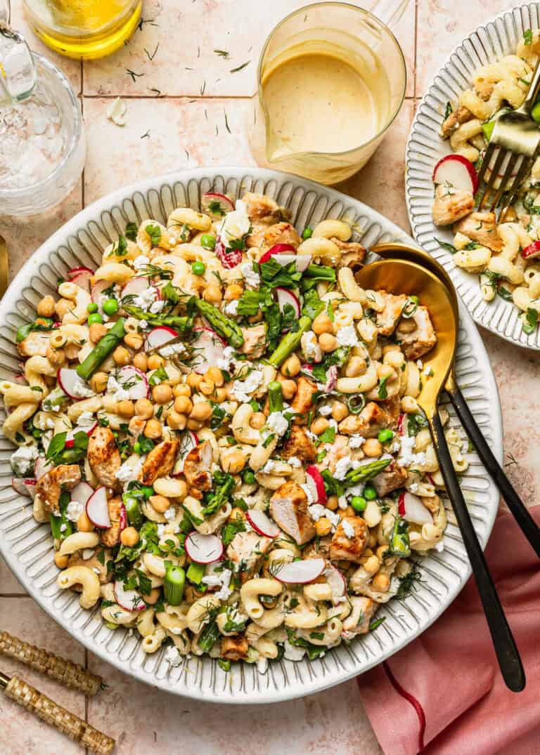 A large bowl of pasta salad with grilled chicken, radish slices, chickpeas, green peas, asparagus, herbs, and feta cheese. Two serving spoons rest in the bowl; a glass of dressing and a pink napkin are nearby.