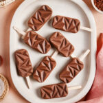 Eight geometric-shaped chocolate popsicles arranged on a white oval plate, set on a peach-colored surface with woven baskets and a pink cloth nearby.