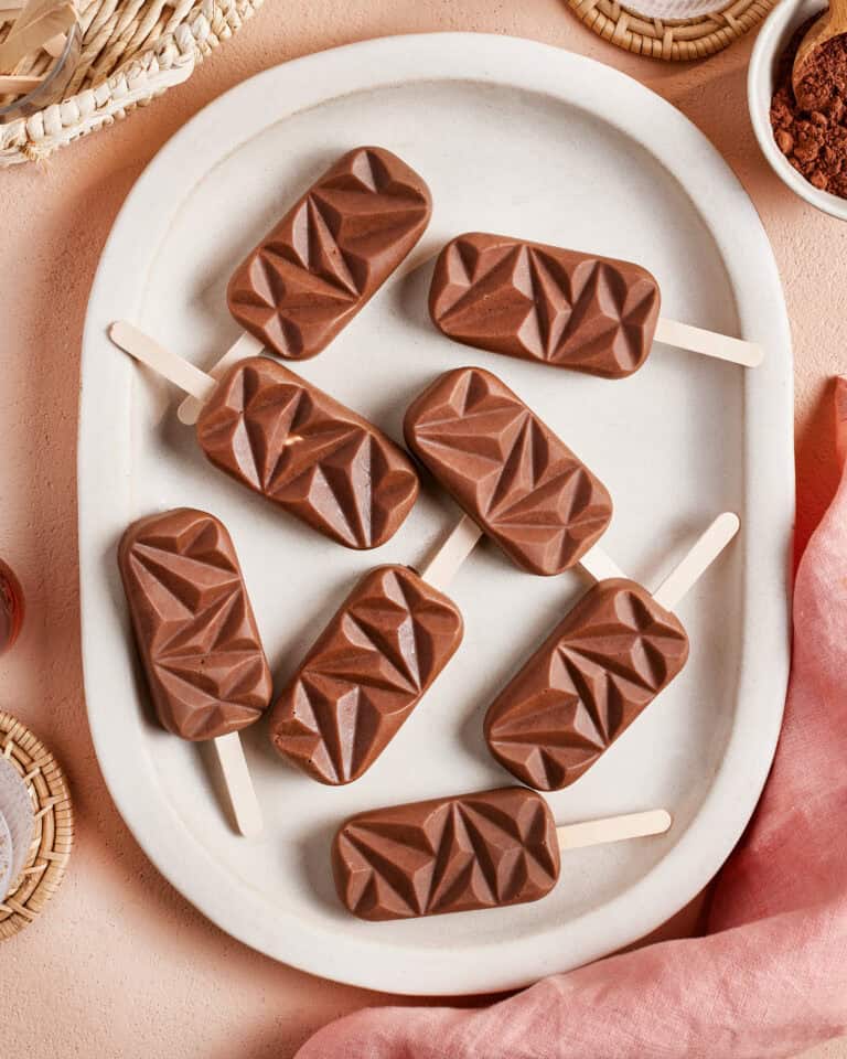 Eight geometric-shaped chocolate popsicles arranged on a white oval plate, set on a peach-colored surface with woven baskets and a pink cloth nearby.