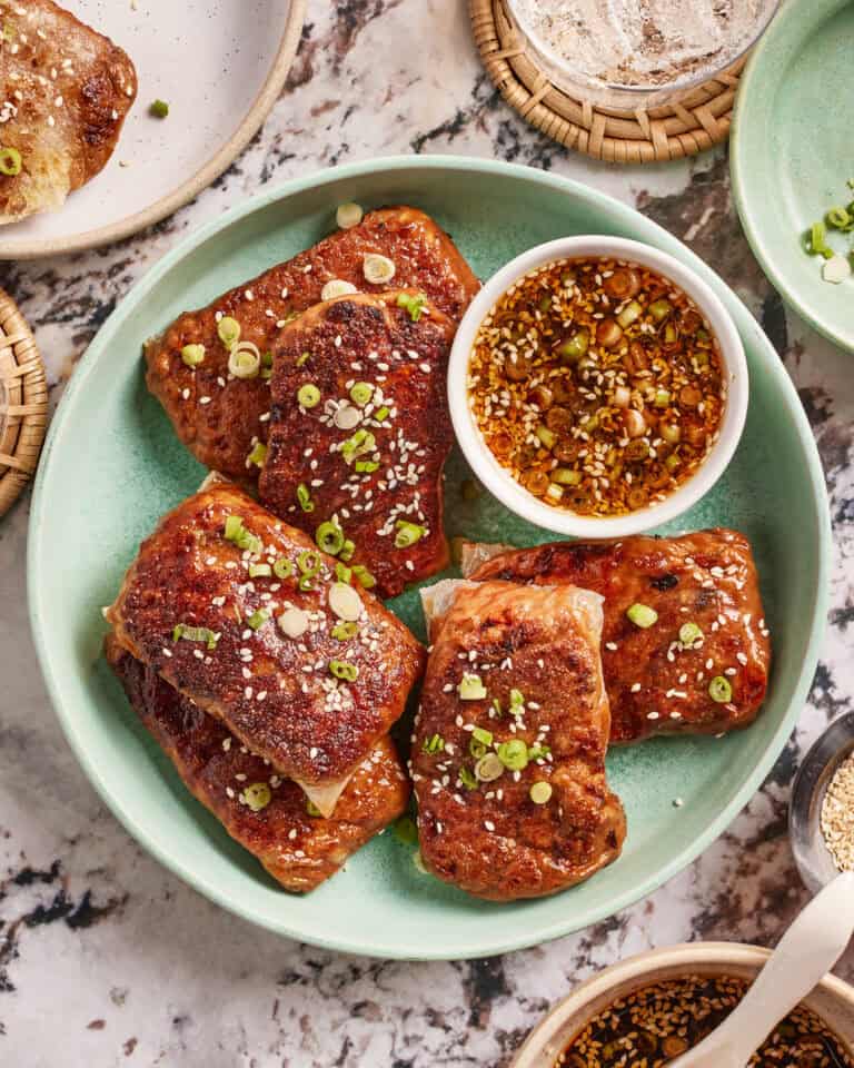 A green plate with five pieces of grilled chicken garnished with sesame seeds and chopped green onions, served with a bowl of dipping sauce on a marble table.