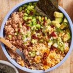 A bowl filled with shredded tuna, edamame, avocado cubes, seaweed sheets, scallions, seasoned rice, and a drizzle of chili oil, with a fork resting inside. The bowl sits on a wooden surface.
