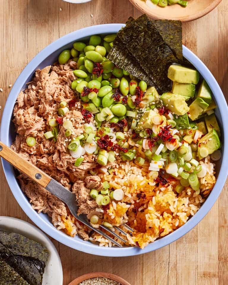 A bowl filled with shredded tuna, edamame, avocado cubes, seaweed sheets, scallions, seasoned rice, and a drizzle of chili oil, with a fork resting inside. The bowl sits on a wooden surface.