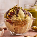 A glass bowl of pistachio ice cream topped with chocolate sauce, chopped pistachios, and shredded pastry, with a jar of pistachio spread and a spoon in the background.