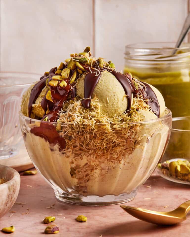 A glass bowl of pistachio ice cream topped with chocolate sauce, chopped pistachios, and shredded pastry, with a jar of pistachio spread and a spoon in the background.