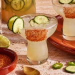 Two margarita glasses filled with a light green drink, ice, and cucumber slices, with chili powder on the rim. A jar of cucumber water, jalapeño slices, a bowl of chili powder, and a spoon are nearby.