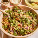 A large wooden bowl filled with a colorful salad of arugula, quinoa, cherry tomatoes, shredded cabbage, corn, and grated cheese. Wooden salad servers rest in the bowl. Plates, forks, and dressing are nearby.