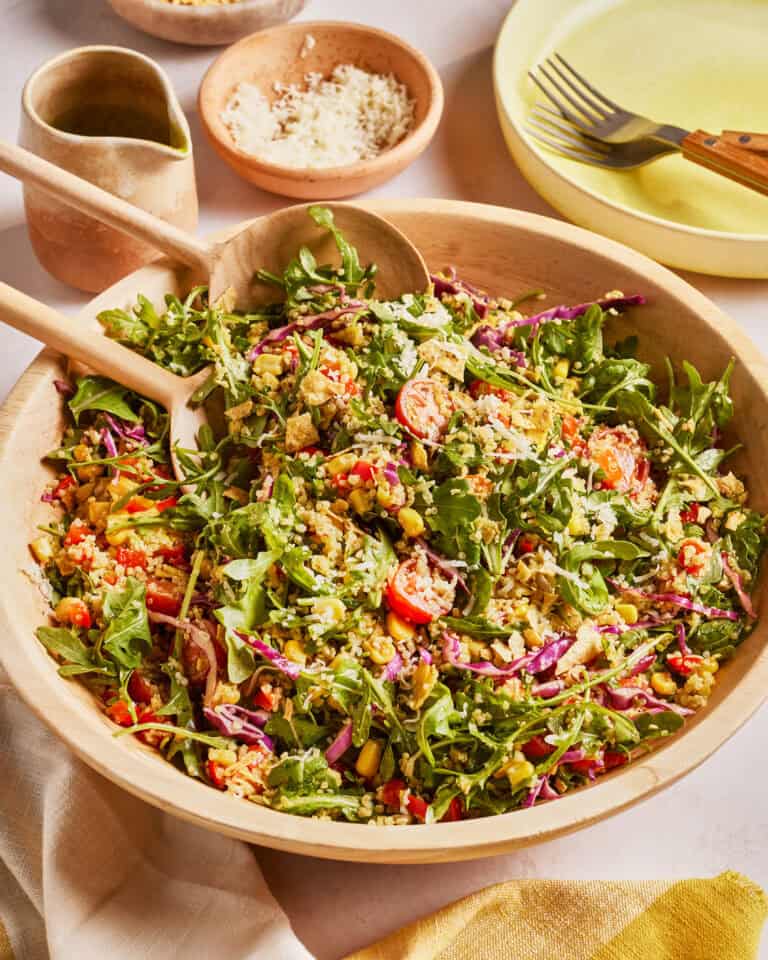A large wooden bowl filled with a colorful salad of arugula, quinoa, cherry tomatoes, shredded cabbage, corn, and grated cheese. Wooden salad servers rest in the bowl. Plates, forks, and dressing are nearby.