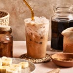 A glass of iced coffee with a gold metal straw sits on a woven coaster, surrounded by jars of coffee, a pitcher, and a plate of sliced bananas on a beige surface.