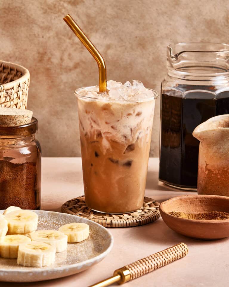A glass of iced coffee with a gold metal straw sits on a woven coaster, surrounded by jars of coffee, a pitcher, and a plate of sliced bananas on a beige surface.
