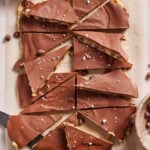 Sliced chocolate-covered bars on parchment paper, sprinkled with flaky sea salt, with two knives and bowls of salt and chocolate chips nearby.