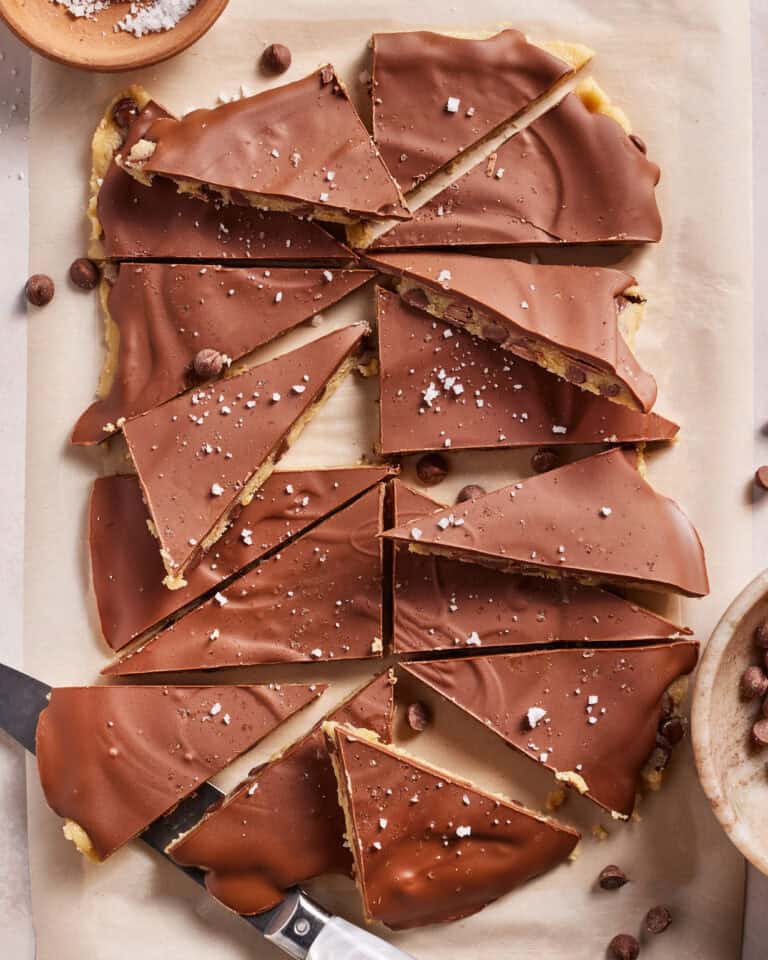 Sliced chocolate-covered bars on parchment paper, sprinkled with flaky sea salt, with two knives and bowls of salt and chocolate chips nearby.