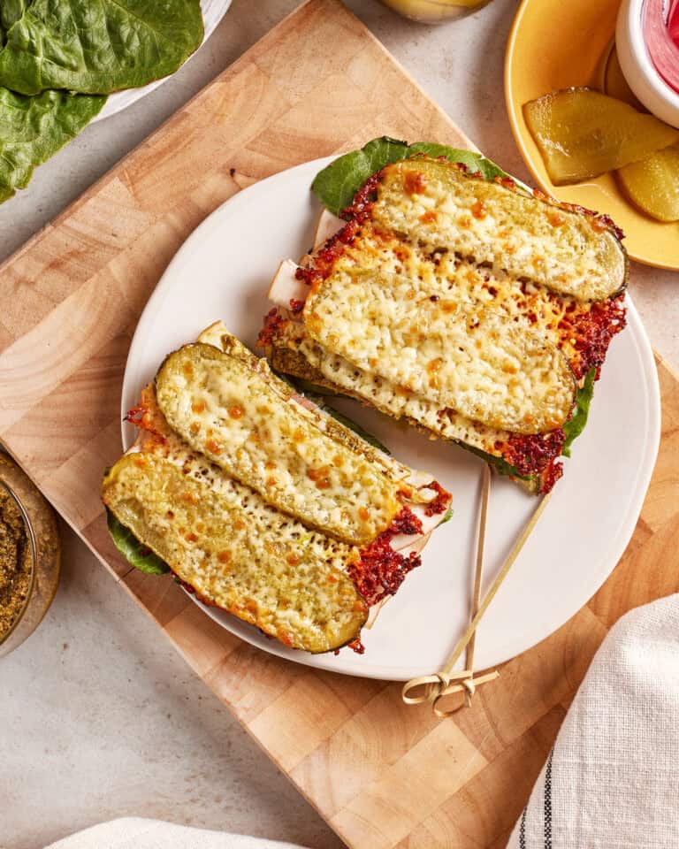 A sandwich cut in half on a white plate, with pickles as the bread, filled with lettuce, cheese, and deli meat. The sandwich is held together with a toothpick and sits on a wooden cutting board.