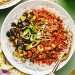 A bowl of white rice topped with spicy red minced tofu, sautéed eggplant, green onions, and sesame seeds, served with a gold fork on a light green table. A striped napkin and another partially visible bowl are nearby.
