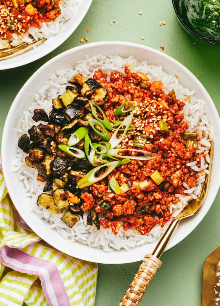 A bowl of white rice topped with spicy red minced tofu, sautéed eggplant, green onions, and sesame seeds, served with a gold fork on a light green table. A striped napkin and another partially visible bowl are nearby.