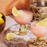 Two frozen pink cocktails garnished with lemon wedges in elegant glasses sit on a terrazzo board, with a bowl of lemons and a decorative pitcher nearby, all in a sunlit setting.