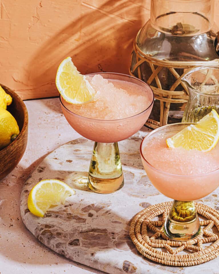 Two frozen pink cocktails garnished with lemon wedges in elegant glasses sit on a terrazzo board, with a bowl of lemons and a decorative pitcher nearby, all in a sunlit setting.