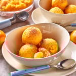 Three scoops of bright orange sorbet topped with grated zest are served in a bowl with a peeled tangerine segment beside a spoon. In the background, more sorbet and an ice cream scoop are visible.