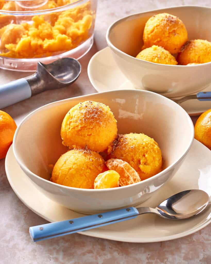 Three scoops of bright orange sorbet topped with grated zest are served in a bowl with a peeled tangerine segment beside a spoon. In the background, more sorbet and an ice cream scoop are visible.