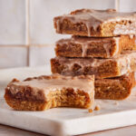 Four frosted blondie bars are stacked on a white marble surface, with the front blondie showing a bite taken out, revealing a soft, moist interior. The frosting is glossy and slightly drips over the edges.