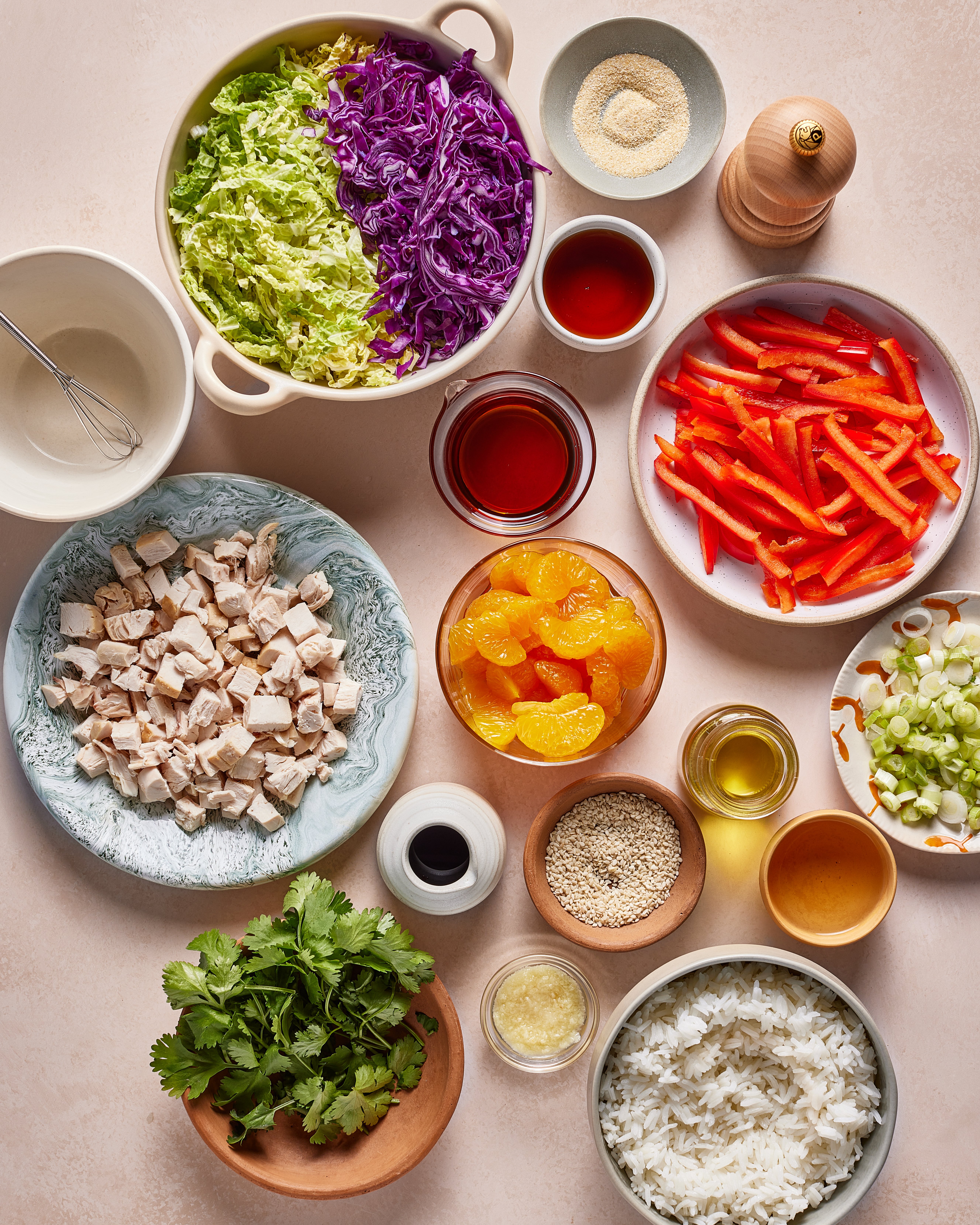 Variety of fresh ingredients in bowls on a table, including shredded cabbage, red bell pepper, cubed chicken, mandarin oranges, cooked rice, green onions, cilantro, sesame seeds, seasonings, and various sauces.