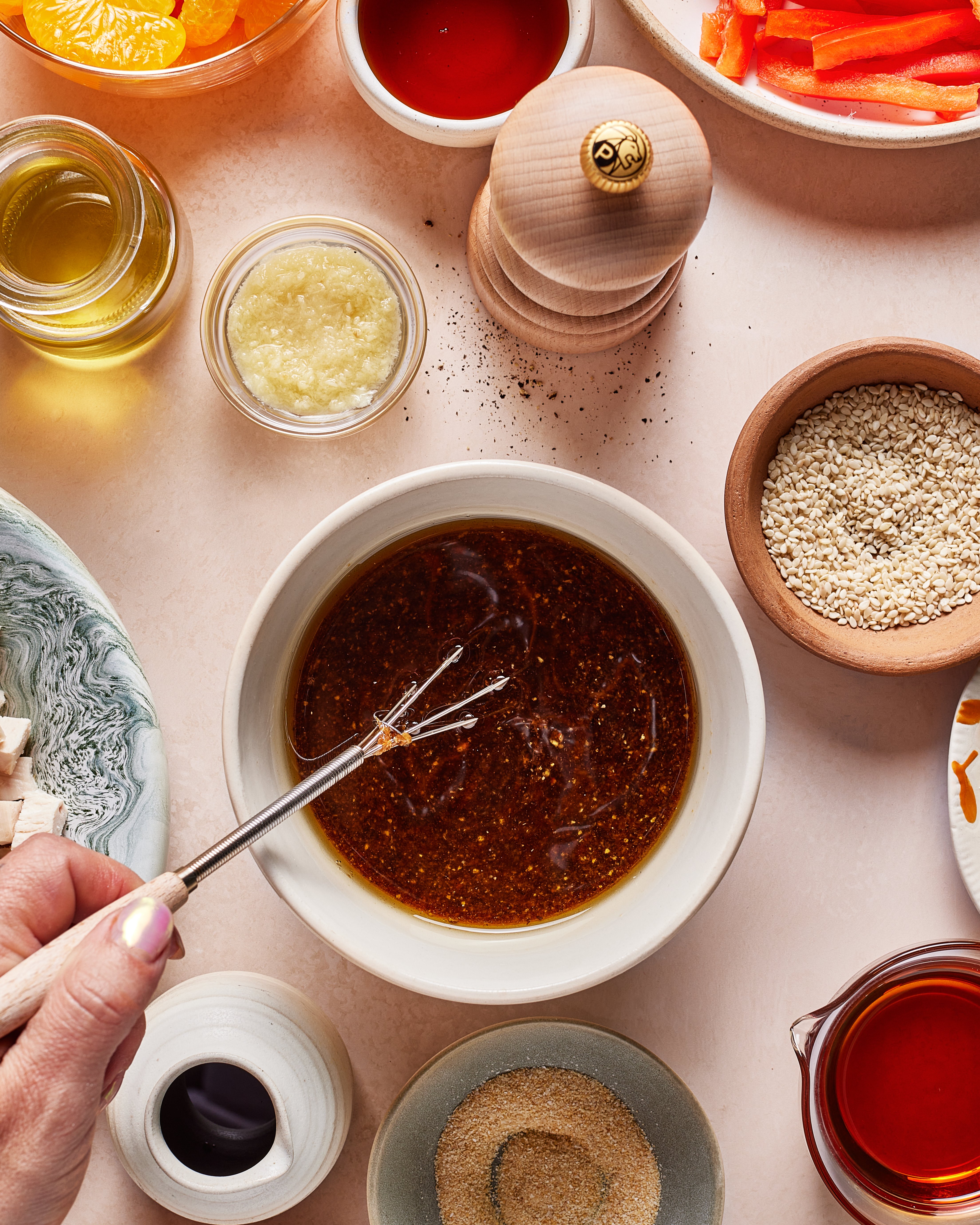 A hand whisks a brown sauce in a white bowl, surrounded by small dishes of oil, minced garlic, pepper, sesame seeds, sliced vegetables, seasonings, and a wooden pepper grinder on a light surface.