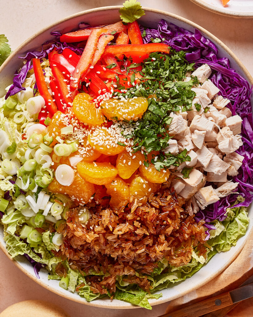 A colorful salad bowl with chopped red bell peppers, diced chicken, shredded purple cabbage, mandarin orange slices, fried onions, chopped green onions, cilantro, and a sprinkle of sesame seeds.