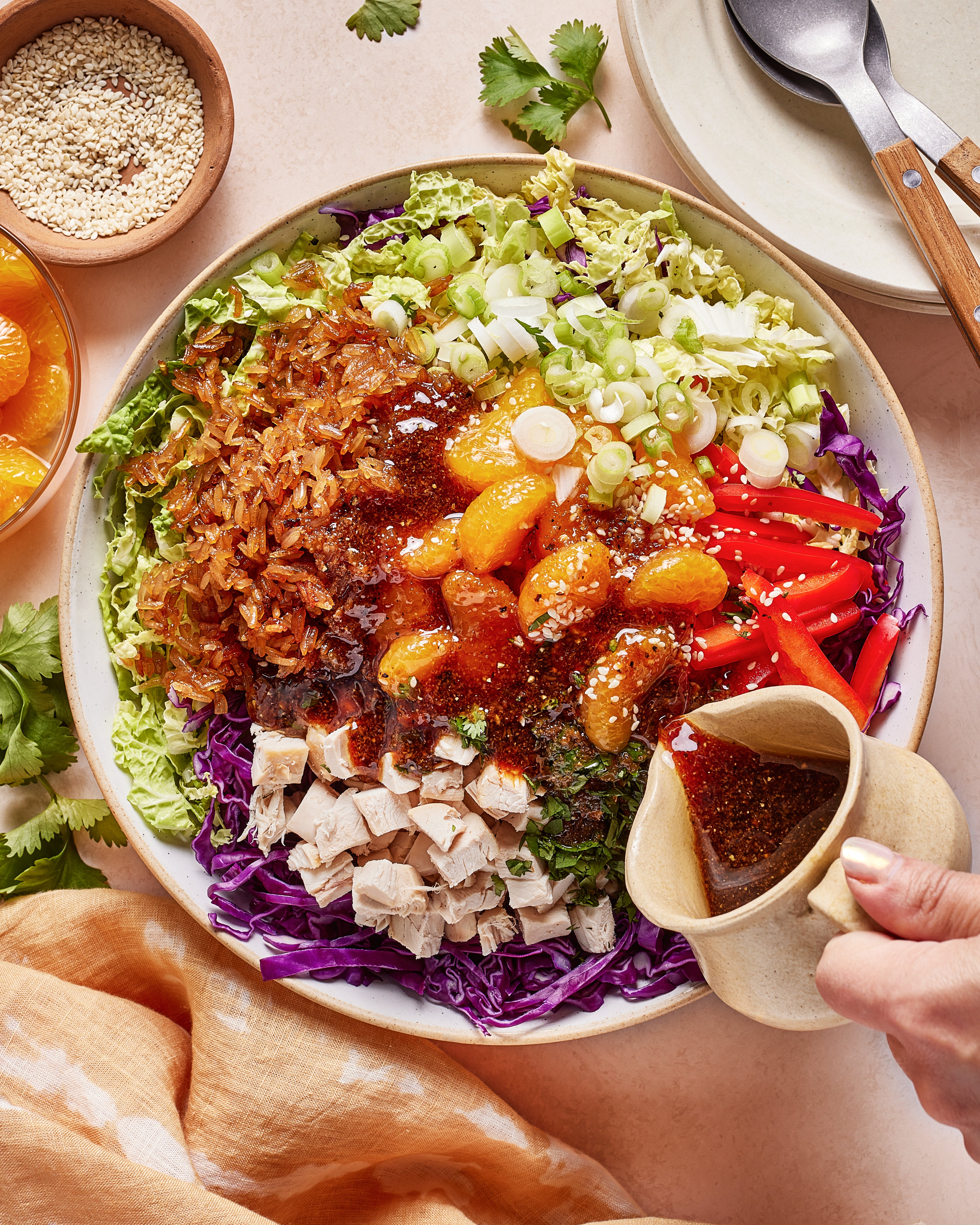 A colorful salad with chopped lettuce, shredded cabbage, red bell pepper, green onions, diced chicken, mandarins, crispy toppings, and sesame seeds is being drizzled with a dark dressing from a small pitcher.