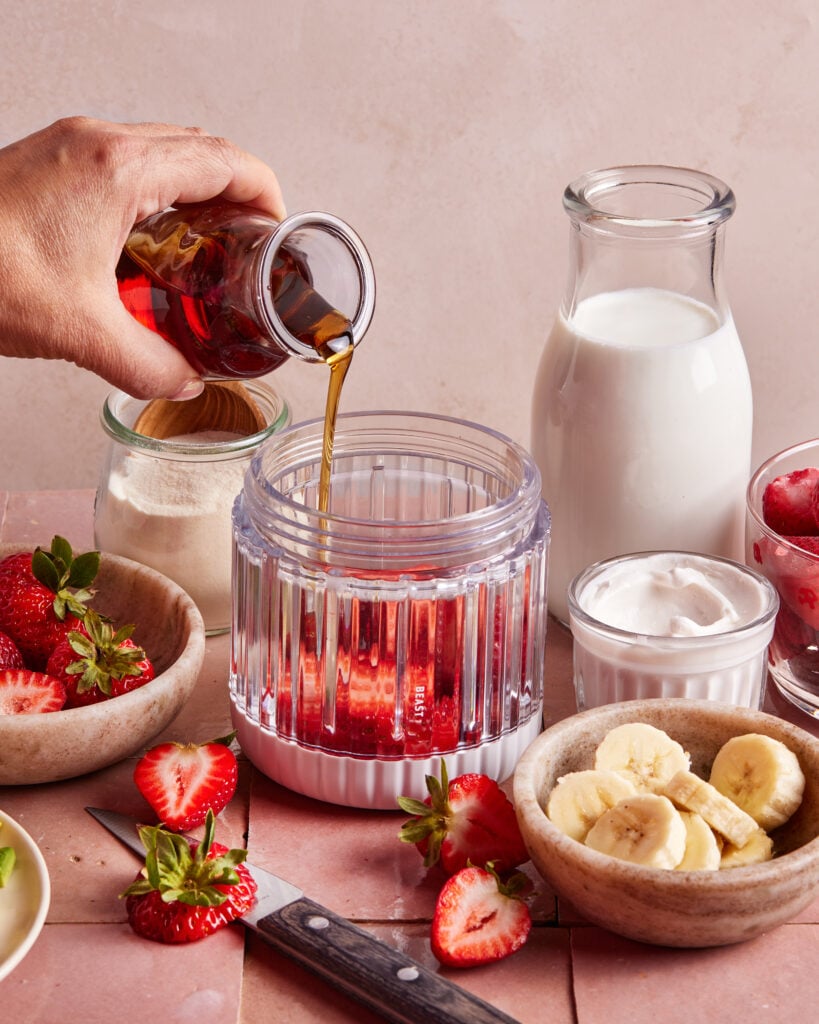 A hand pours a brown liquid, possibly syrup or coffee, into a blender cup with strawberries. Surrounding items include milk, yogurt, protein powder, sliced bananas, and whole strawberries on a light pink surface.