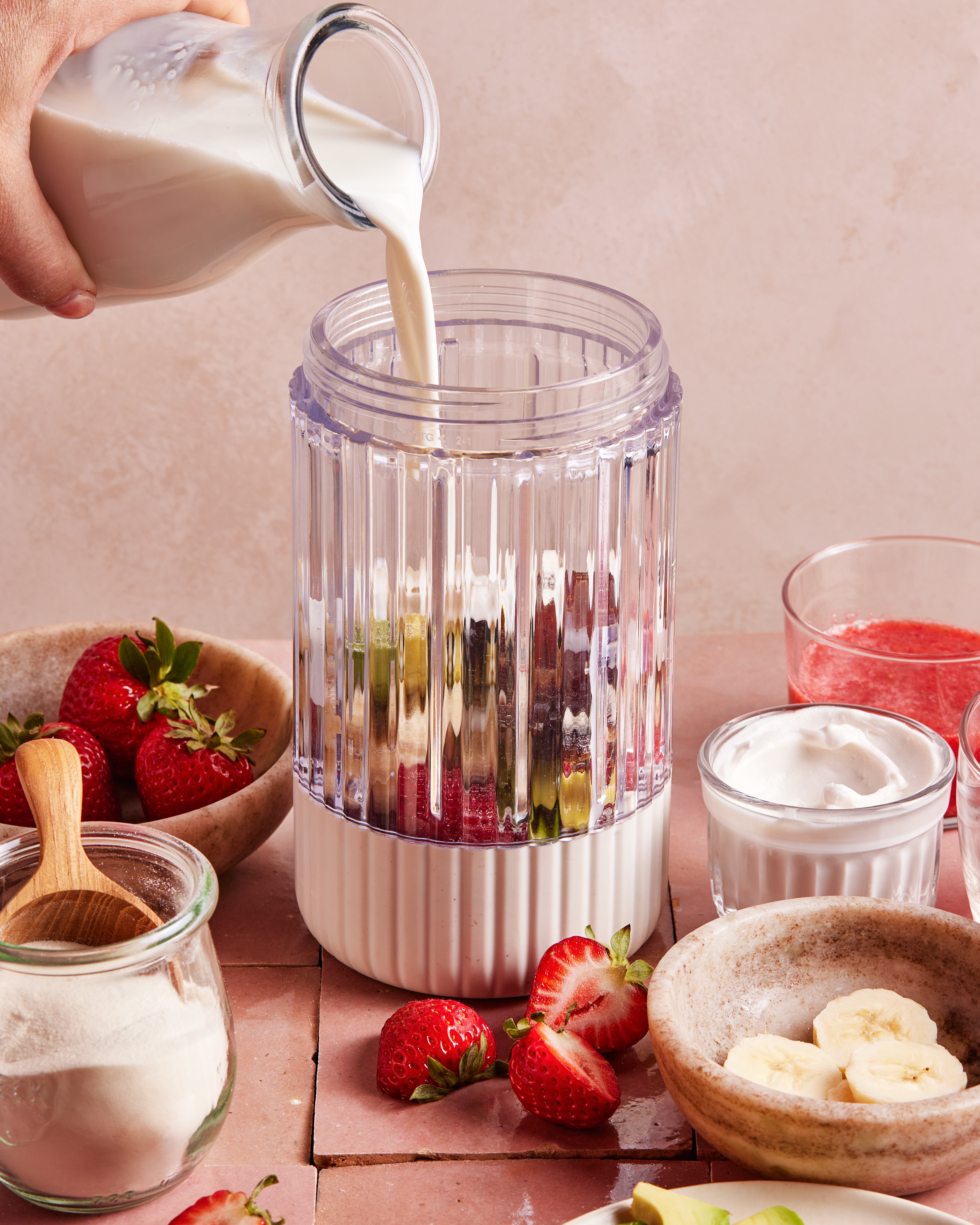 A hand pours milk into a blender filled with cut fruit, surrounded by bowls of strawberries, banana slices, yogurt, and a glass of pink smoothie on a pink surface.