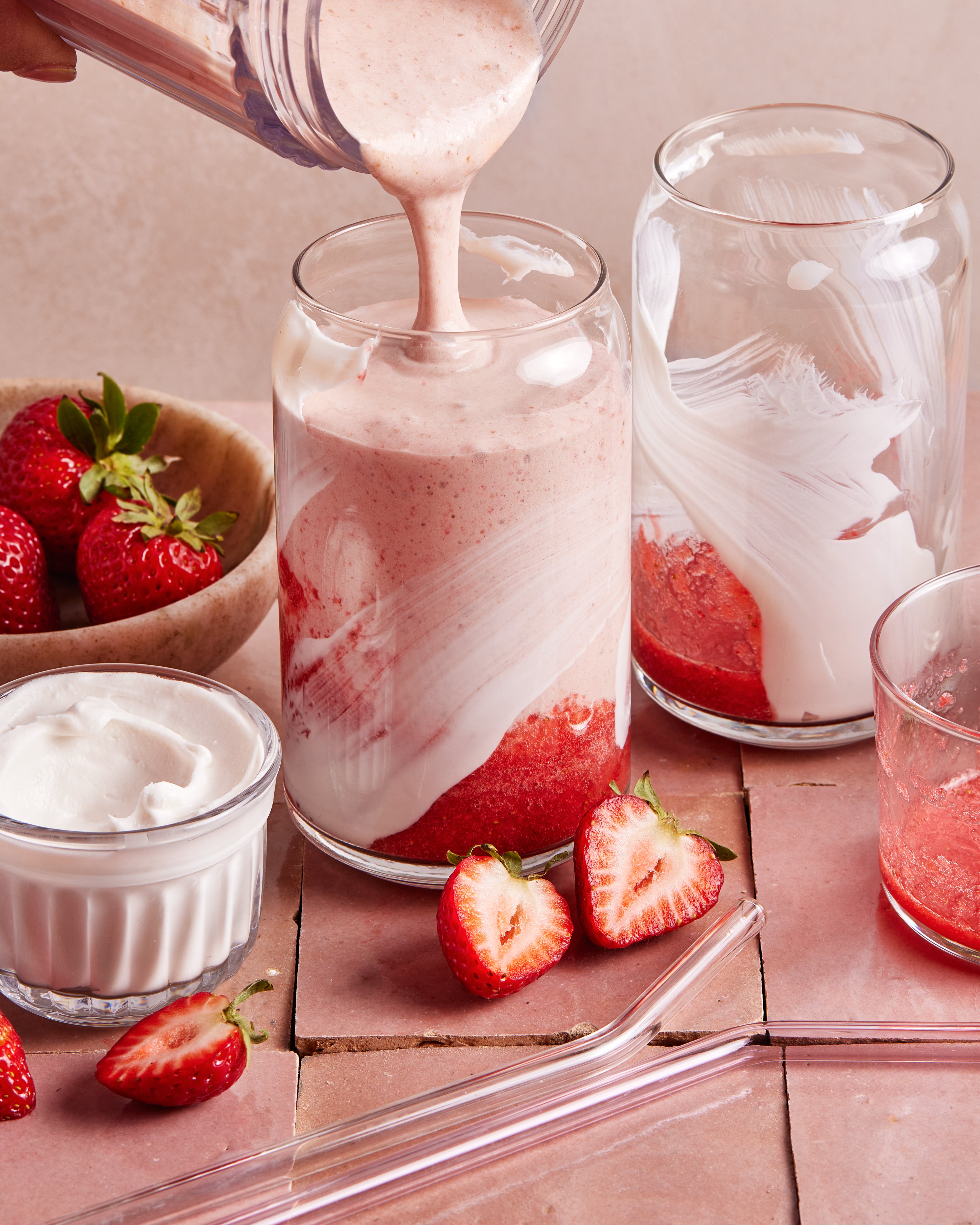 A creamy strawberry smoothie is being poured into a glass with strawberry puree and yogurt, surrounded by fresh strawberries, a glass straw, and a small bowl of yogurt on a pink tiled surface.