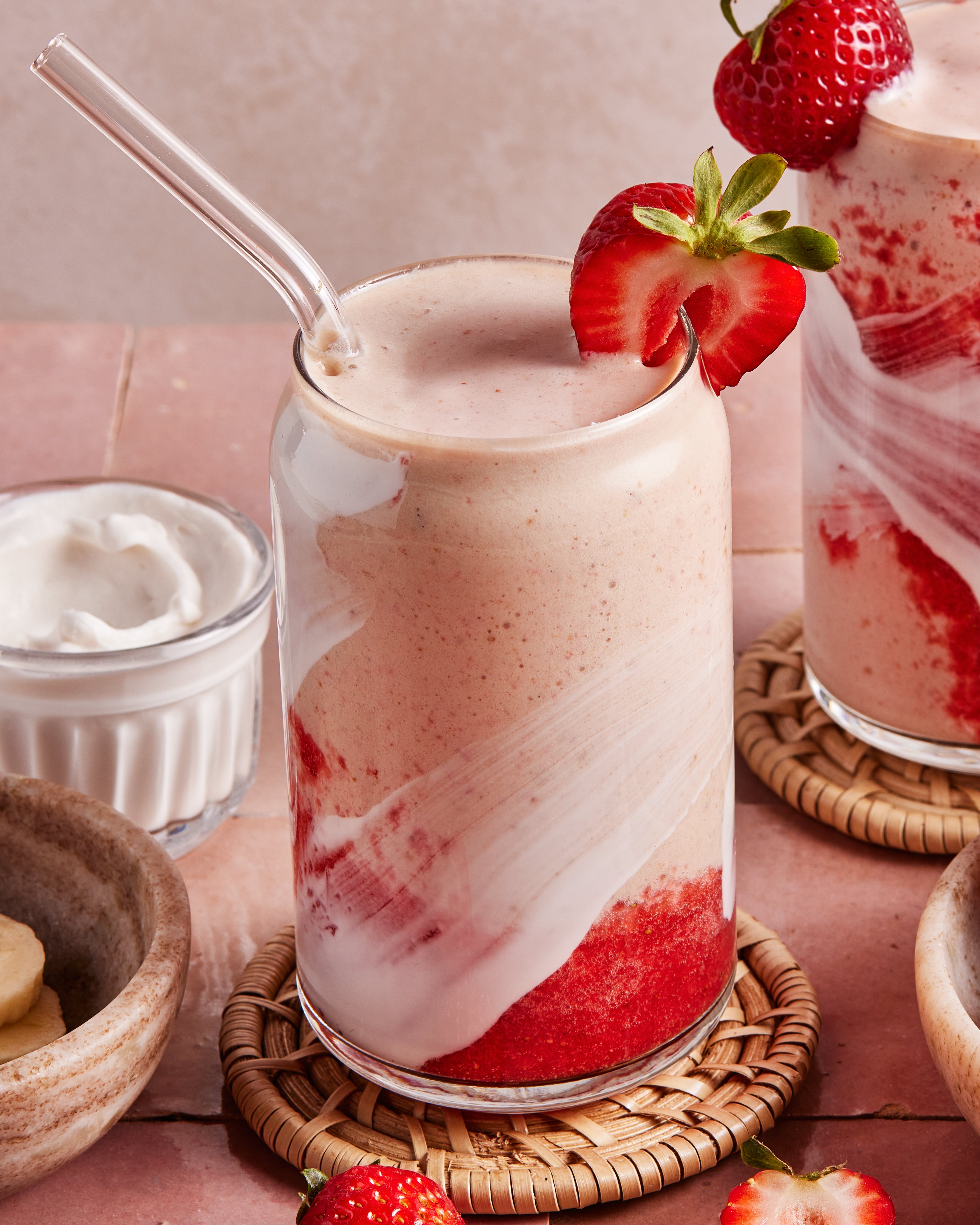 A glass of strawberry smoothie with swirls of yogurt, topped with a sliced strawberry and served with a glass straw. Nearby are fresh strawberries, banana slices, and a small bowl of whipped cream.