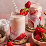 Two glasses of strawberry and cream smoothie with swirled red and white layers, topped with fresh strawberries and clear straws. Fresh strawberries and a bowl of cream are nearby on a pink tiled surface.