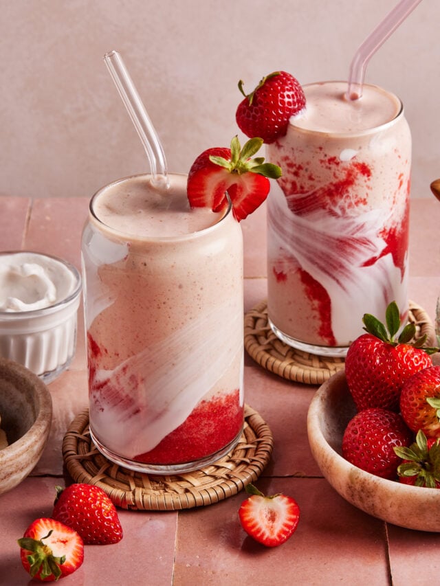 Two glasses of strawberry and cream smoothie with swirled red and white layers, topped with fresh strawberries and clear straws. Fresh strawberries and a bowl of cream are nearby on a pink tiled surface.