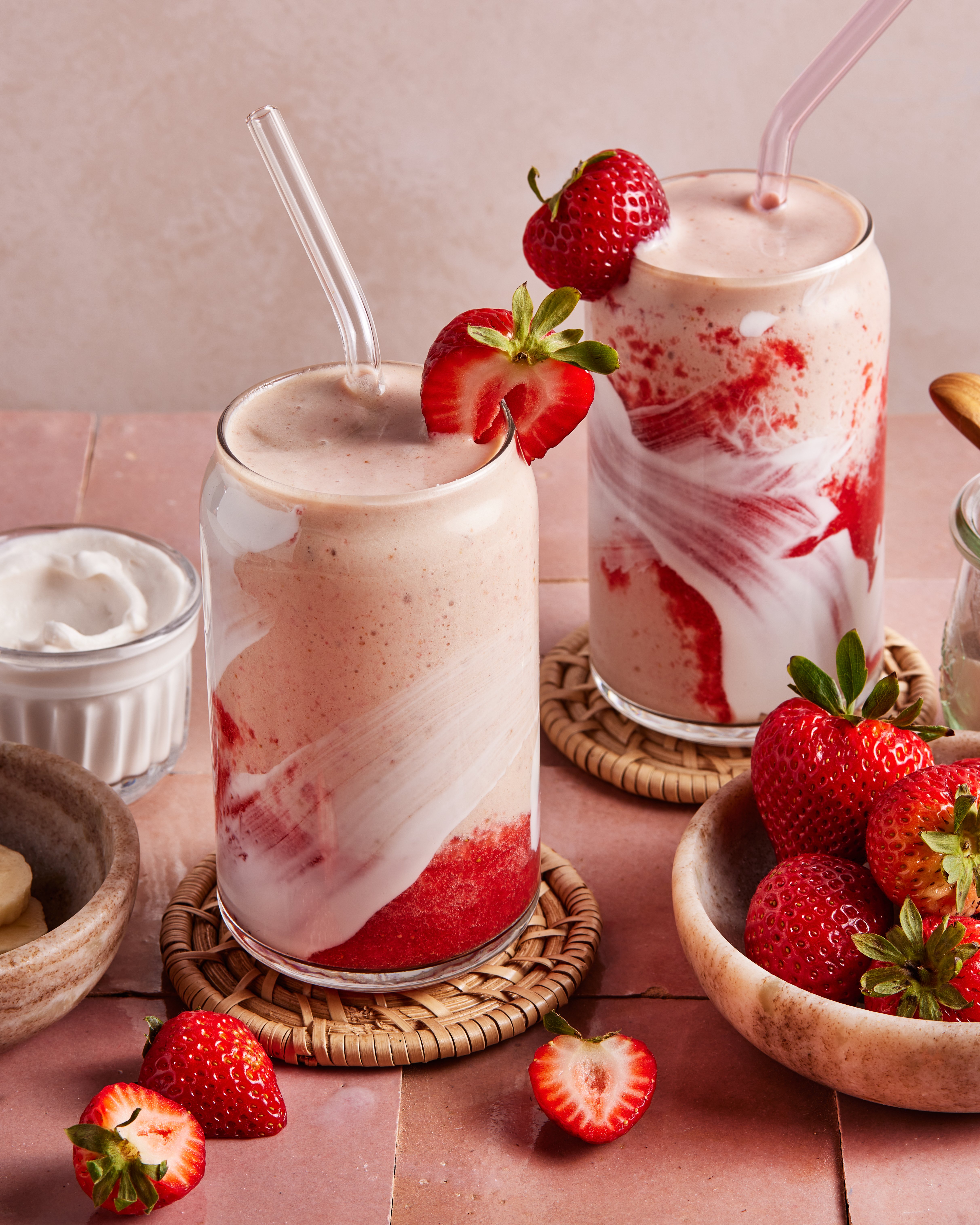 Two glasses of strawberry and cream smoothie with swirled red and white layers, topped with fresh strawberries and clear straws. Fresh strawberries and a bowl of cream are nearby on a pink tiled surface.