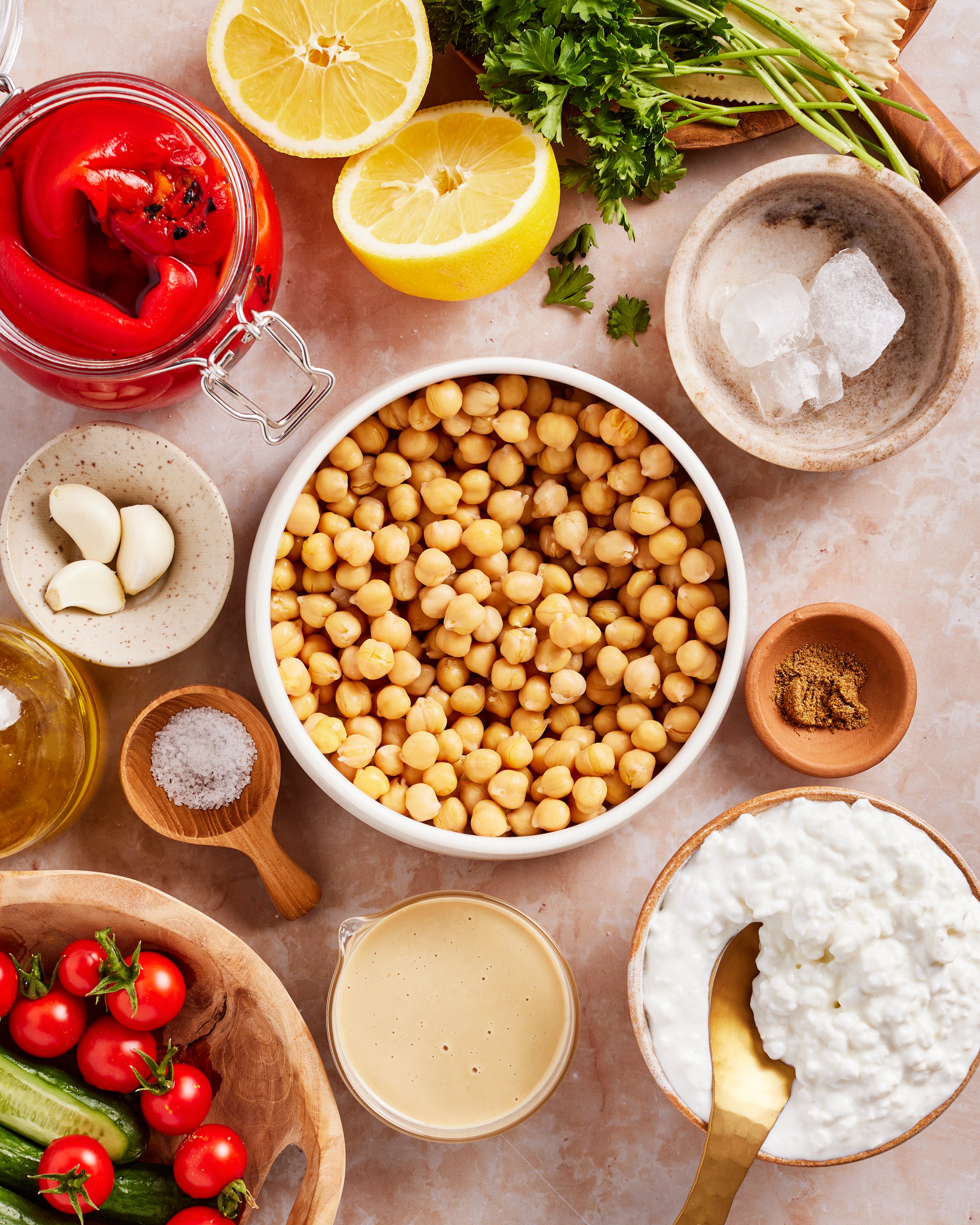 A bowl of chickpeas surrounded by ingredients like lemon halves, roasted red peppers, fresh parsley, cottage cheese, garlic, grape tomatoes, cucumber, olive oil, spices, and coarse salt on a marble surface.
